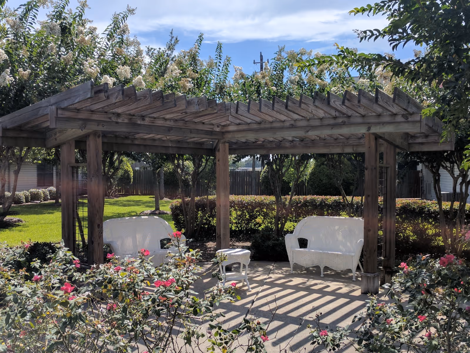 Wooden pergola over a patio with white wicker chairs and a small table surrounded by flowering shrubs and a lawn.
