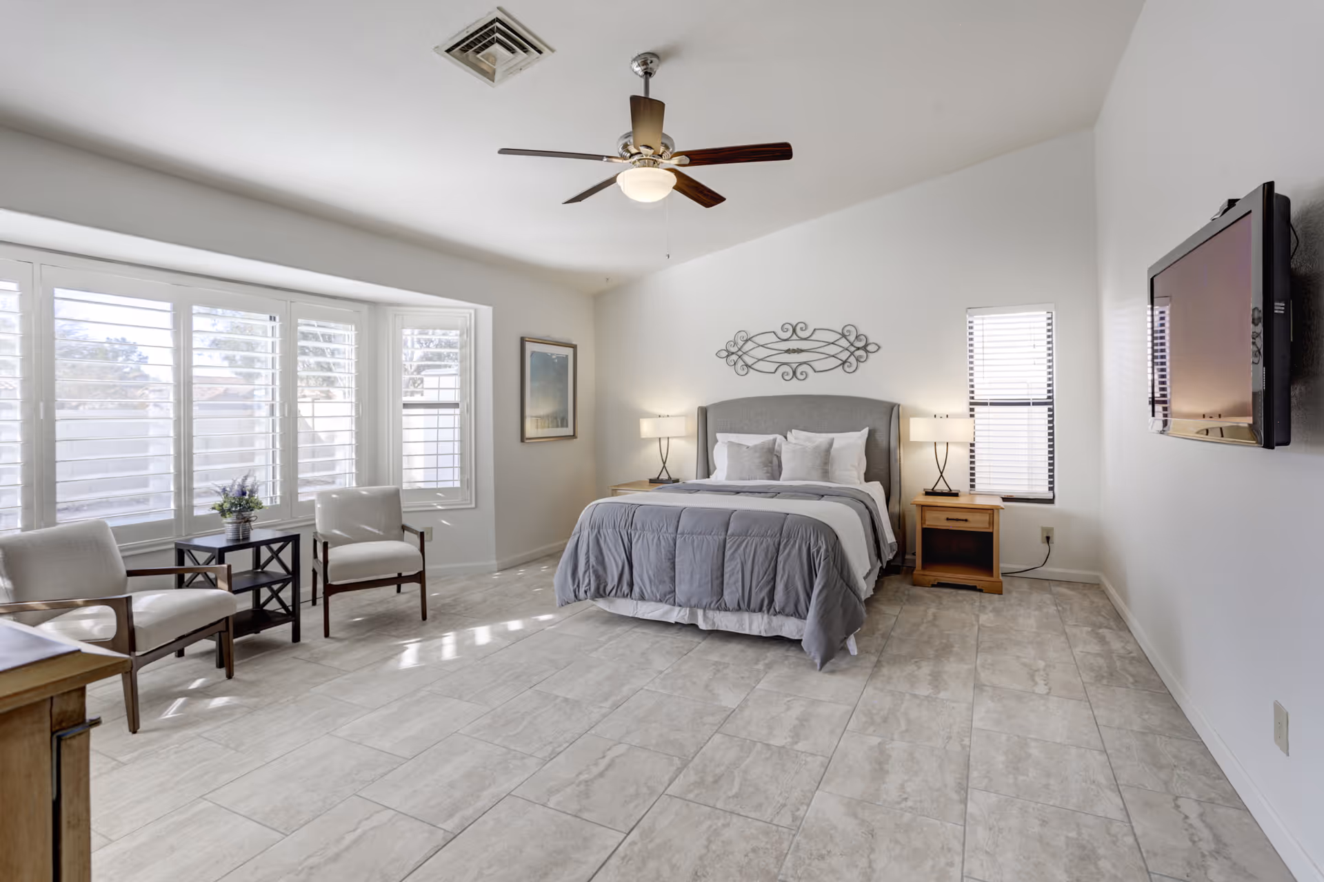 A bright and spacious bedroom with a large bed centered against the wall, flanked by two wooden nightstands with lamps. The room features a ceiling fan, two armchairs near a window with white shutters, a small table with a flower vase, a wall-mounted flat-screen TV, and light-colored tiled flooring.