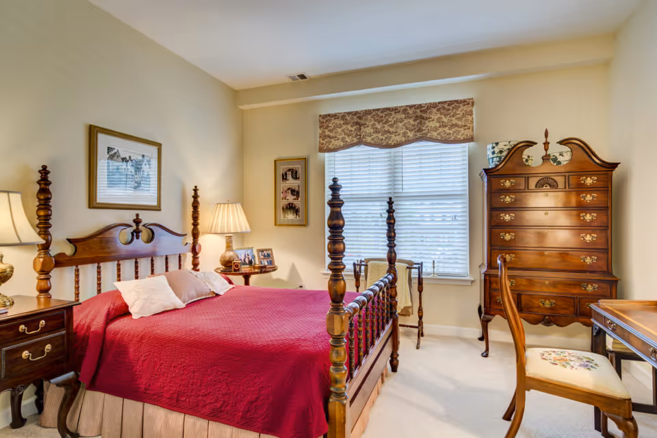 A cozy bedroom with a wooden four-poster bed covered with a red quilt and pillows. There are two bedside tables with lamps, a wooden dresser with decorative items on top, a wooden chair with a floral cushion, and a window with white blinds and a patterned valance. The walls are light-colored and decorated with framed pictures.