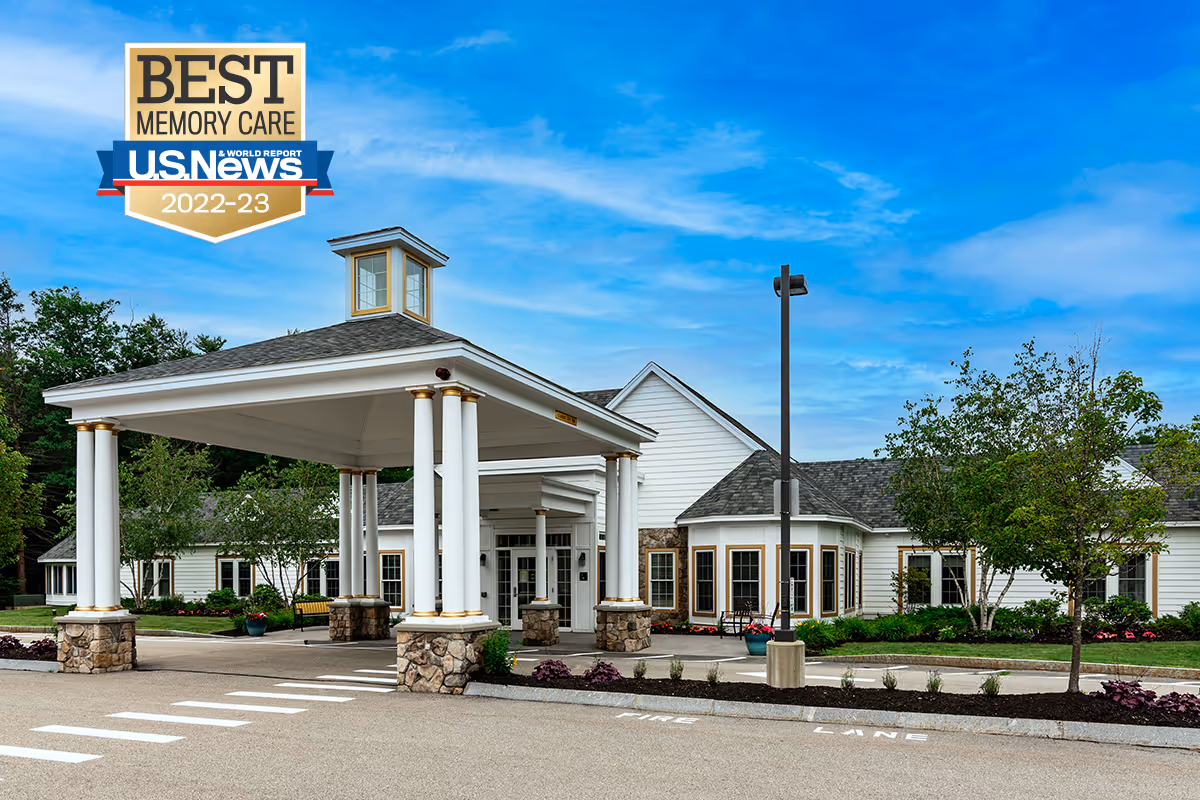 Exterior view of Benchmark at Rye senior living facility showing a covered entrance with white columns and stone bases, surrounded by landscaped greenery and trees under a blue sky.