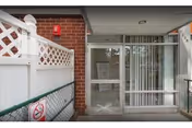 Entrance area of a building with a glass door and vertical blinds on the right side. There is a white fence with lattice design on the left and a brick wall behind it. A no smoking sign is visible on the fence.