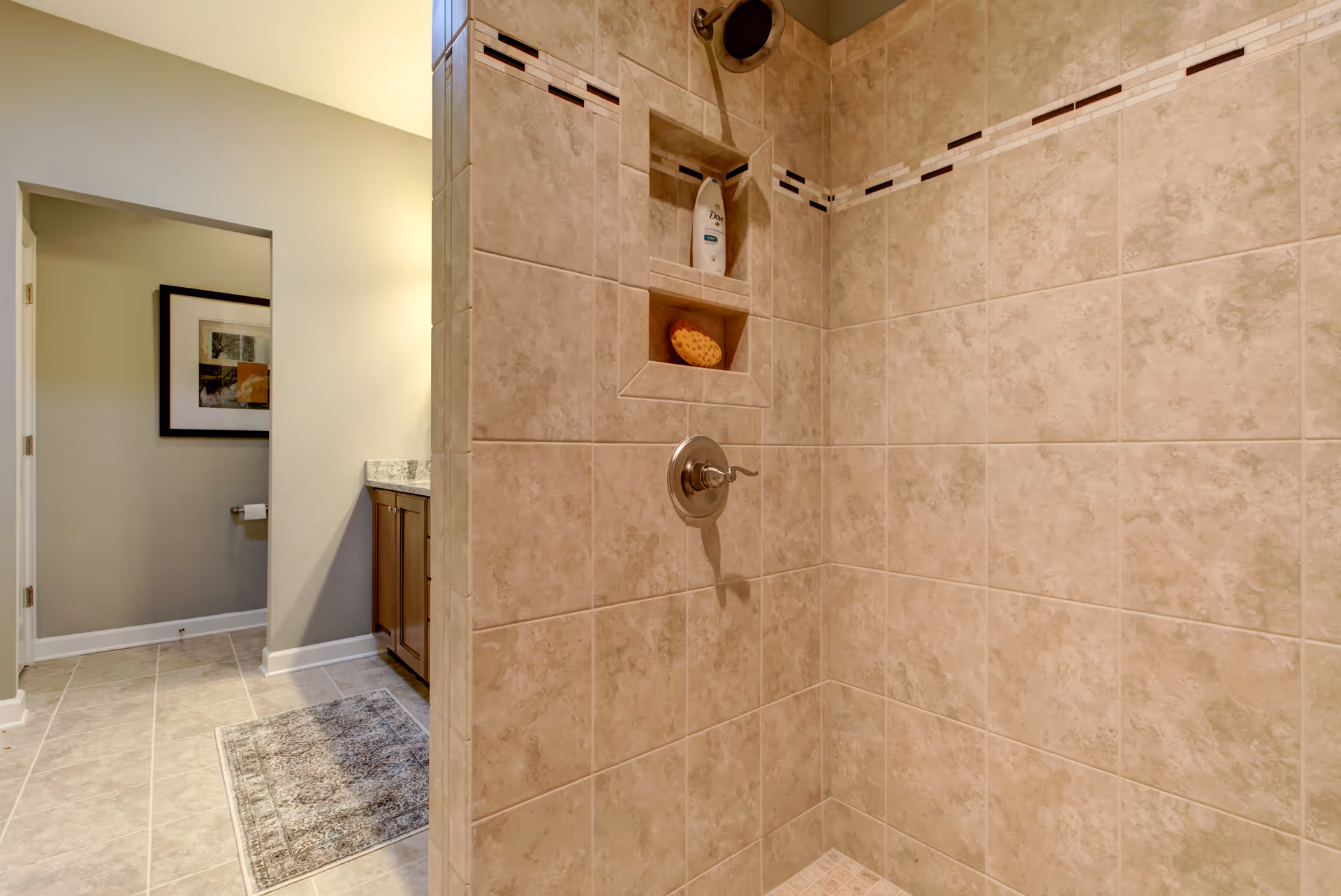 A tiled shower area with beige tiles and a built-in niche holding a bottle of shampoo and a sponge. The shower has a silver handle and a round showerhead. In the background, there is a bathroom vanity with a granite countertop, a framed picture on the wall, and a toilet paper holder.