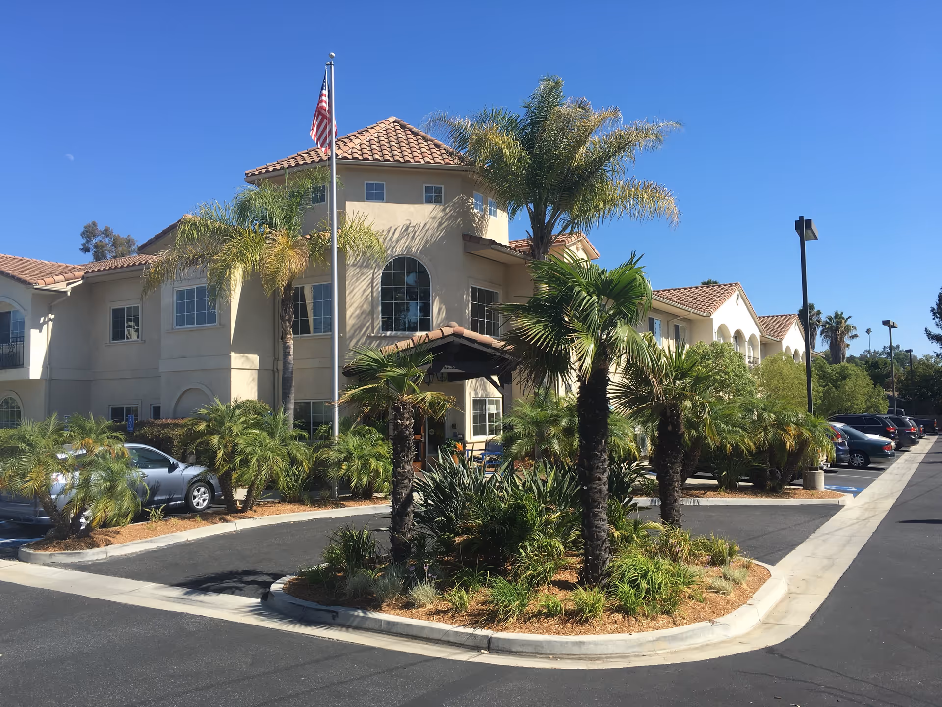Exterior view of a two-story building with a tiled roof and multiple windows, surrounded by palm trees and other greenery. There is an American flag on a flagpole in front of the building, and a parking lot with several cars is visible on the right side under a clear blue sky.