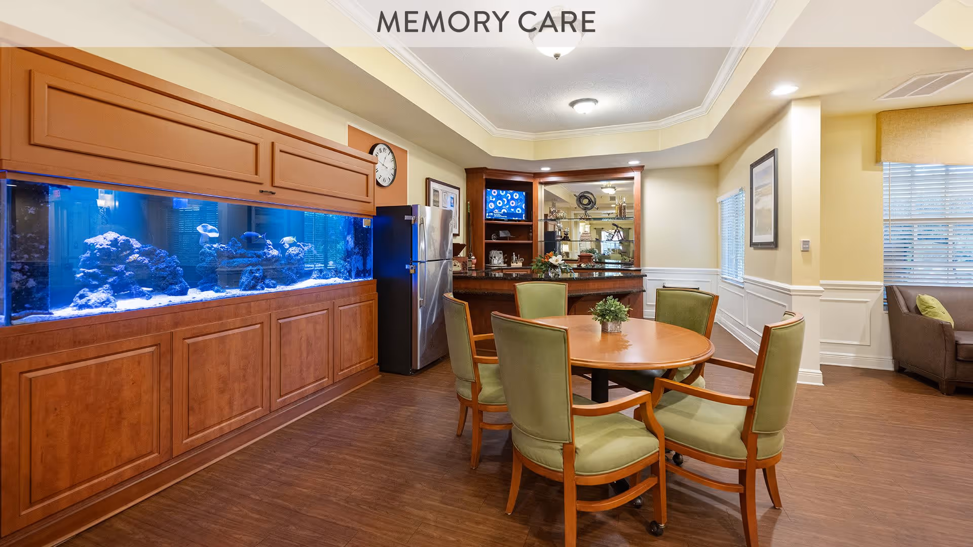 Interior view of a memory care common area with a large built-in aquarium on the left, a round wooden table surrounded by six green cushioned chairs in the center, and a small bar area with a refrigerator and shelves in the background. The room has wood flooring, beige walls with white wainscoting, and windows with blinds allowing natural light.