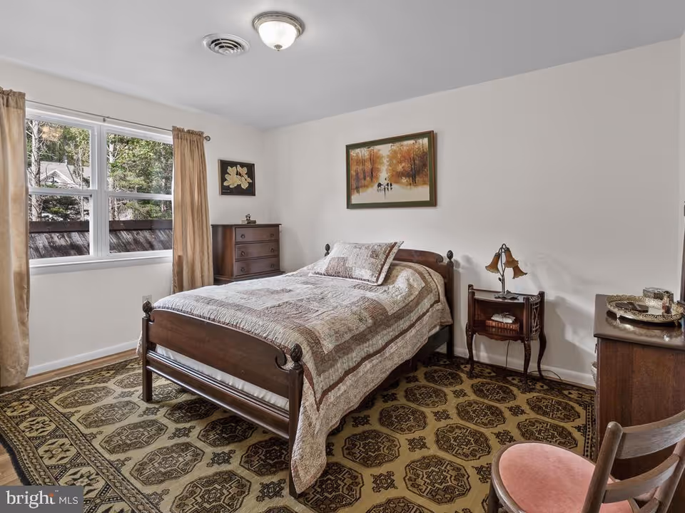 A bedroom with a wooden bed frame and a quilted bedspread. There is a patterned carpet on the floor, a wooden dresser, a small side table with a vintage lamp, and a wooden chair with a pink cushion. Two framed pictures hang on the white walls, and a window with beige curtains lets in natural light.