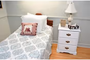 A neatly made bedroom with a patterned bed, white nightstand, lamp, and pastel accents.