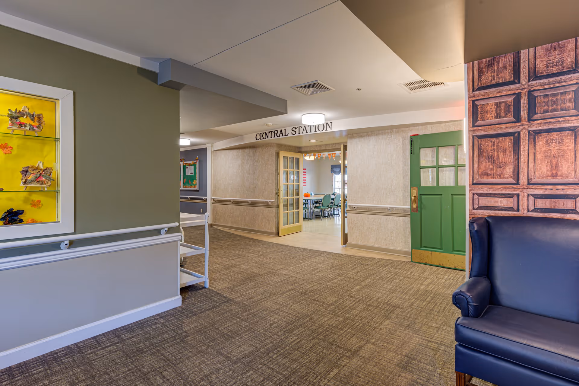 Interior hallway of a senior living facility with beige walls and carpeted floor. On the right side, there is a dark blue armchair and a wall with a wood panel design. Ahead, double glass doors labeled 'Central Station' lead to a room with tables and chairs. A green door is partially open on the right side of the hallway. On the left wall, there is a display case with autumn-themed decorations.