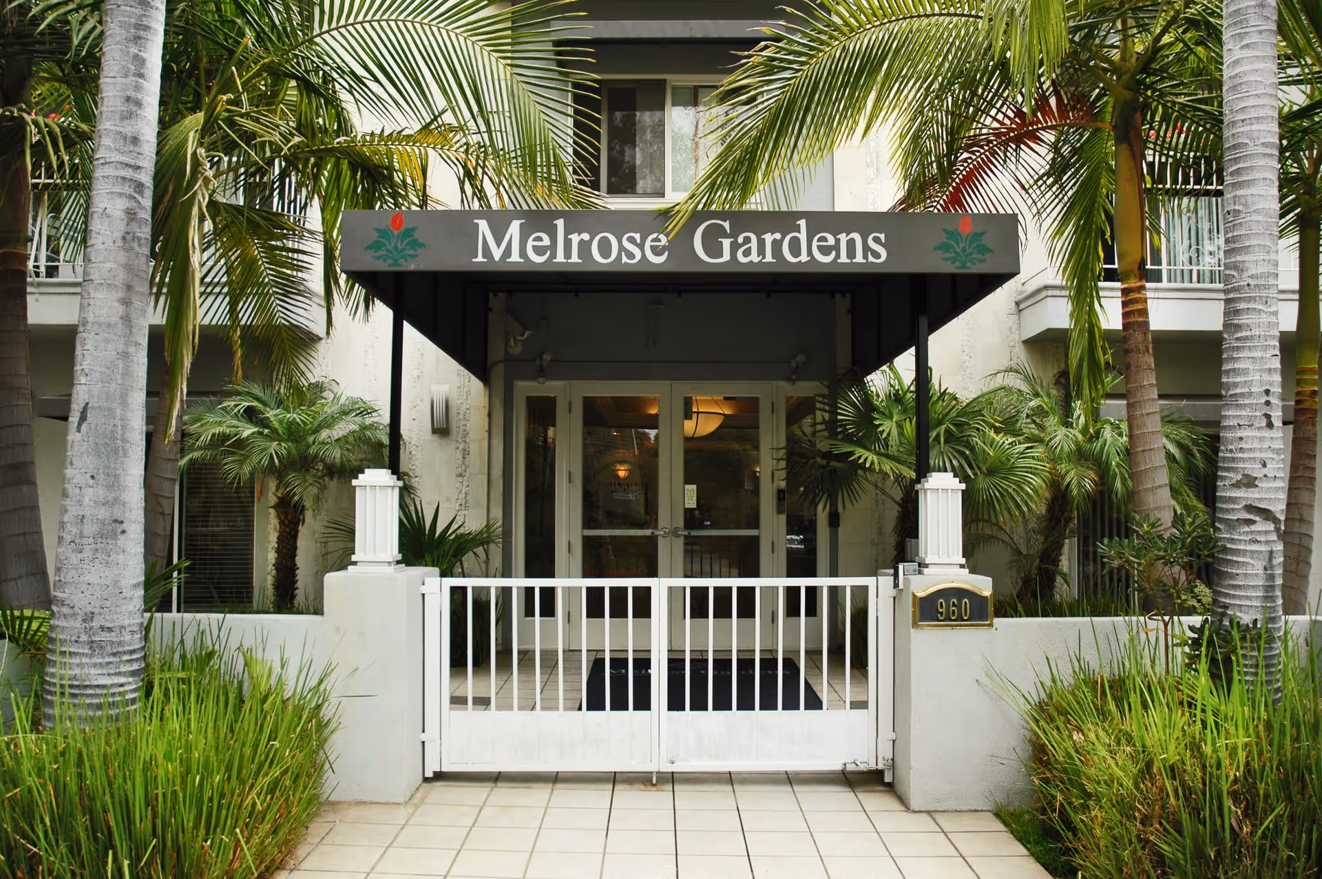 Entrance to Melrose Gardens facility with a white gate, tiled walkway, and a canopy sign displaying the name Melrose Gardens. The entrance is flanked by palm trees and lush greenery.