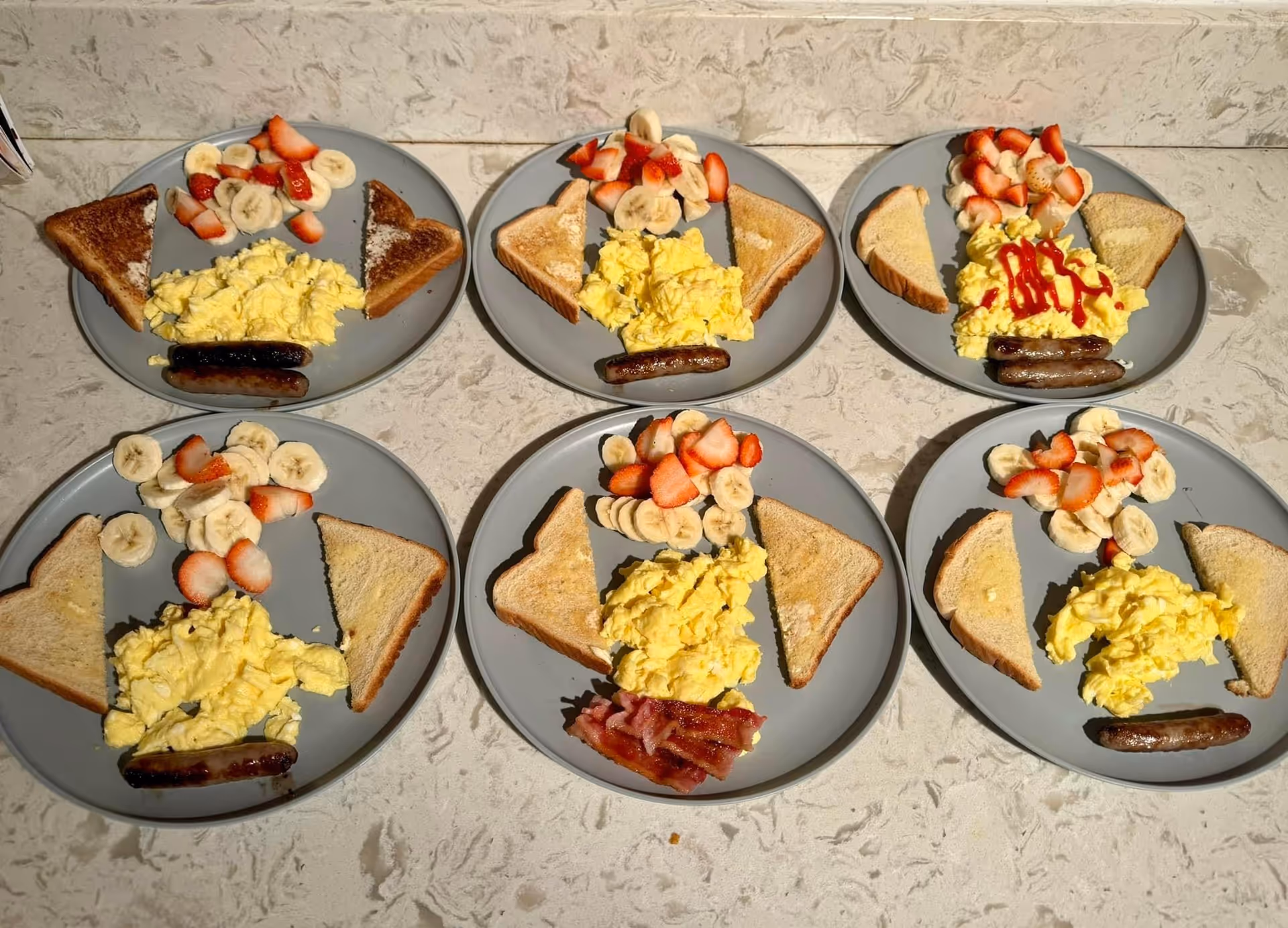 Six plates of breakfast arranged on a countertop, each plate containing scrambled eggs, two slices of toasted bread, sliced bananas and strawberries, and either a sausage link or bacon. One plate has scrambled eggs topped with ketchup.
