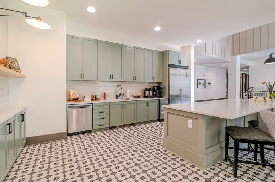 A modern kitchen area with light green cabinets, a large island with a white countertop, patterned black and white tile flooring, stainless steel dishwasher and refrigerator, and recessed ceiling lights.