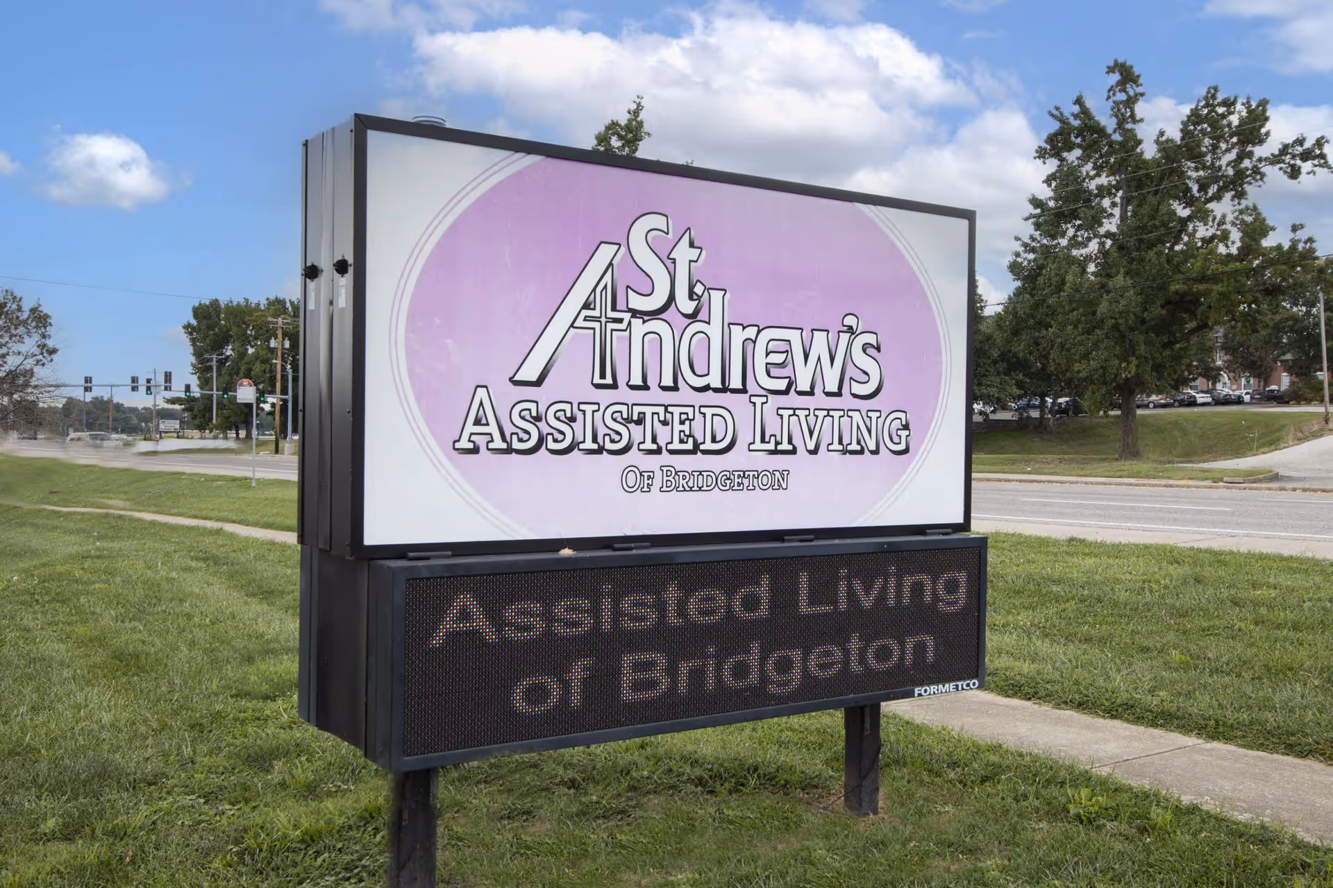 Outdoor sign for St. Andrew's Assisted Living of Bridgeton on a grassy area near a road with trees and a sidewalk in the background under a partly cloudy sky.