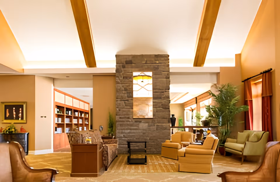 A spacious and warmly lit living room area with wooden ceiling beams, a central stone fireplace, comfortable armchairs and sofas arranged around the fireplace, large windows with red curtains, and indoor plants adding greenery to the space.