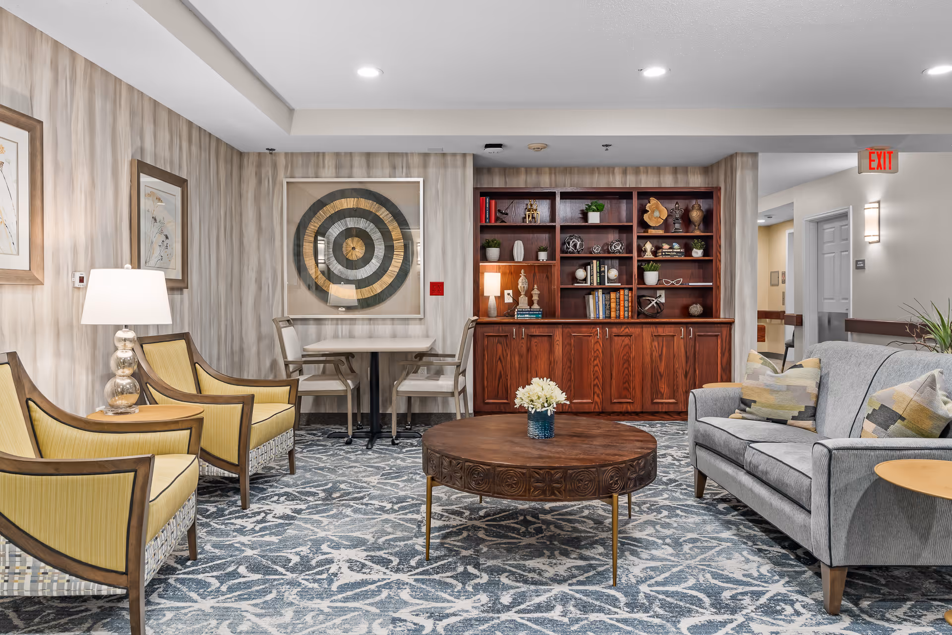 A cozy living room area in Avalon Square featuring a gray sofa with patterned pillows, two yellow armchairs, a round wooden coffee table with a small flower arrangement, a wooden bookshelf with decorative items and books, a small table with two chairs, and framed artwork on the walls. The room has patterned carpet flooring and soft lighting.