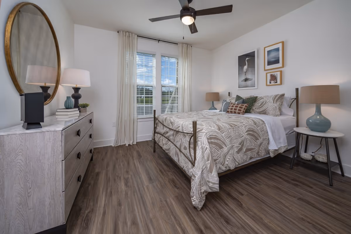 A bright and modern bedroom featuring a bed with patterned bedding and multiple pillows, two bedside tables each with a lamp, a dresser with a round mirror above it, and a window with white curtains letting in natural light. The room has wood flooring and a ceiling fan with a light.