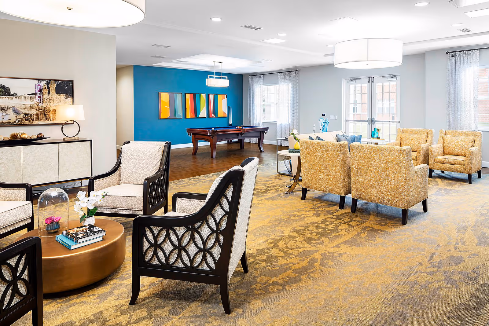 A bright and spacious living room area in a senior living facility featuring multiple seating arrangements with beige and yellow upholstered chairs, a round coffee table with books and flowers, a pool table against a blue accent wall with colorful abstract paintings, large windows with sheer curtains, and modern ceiling lights.