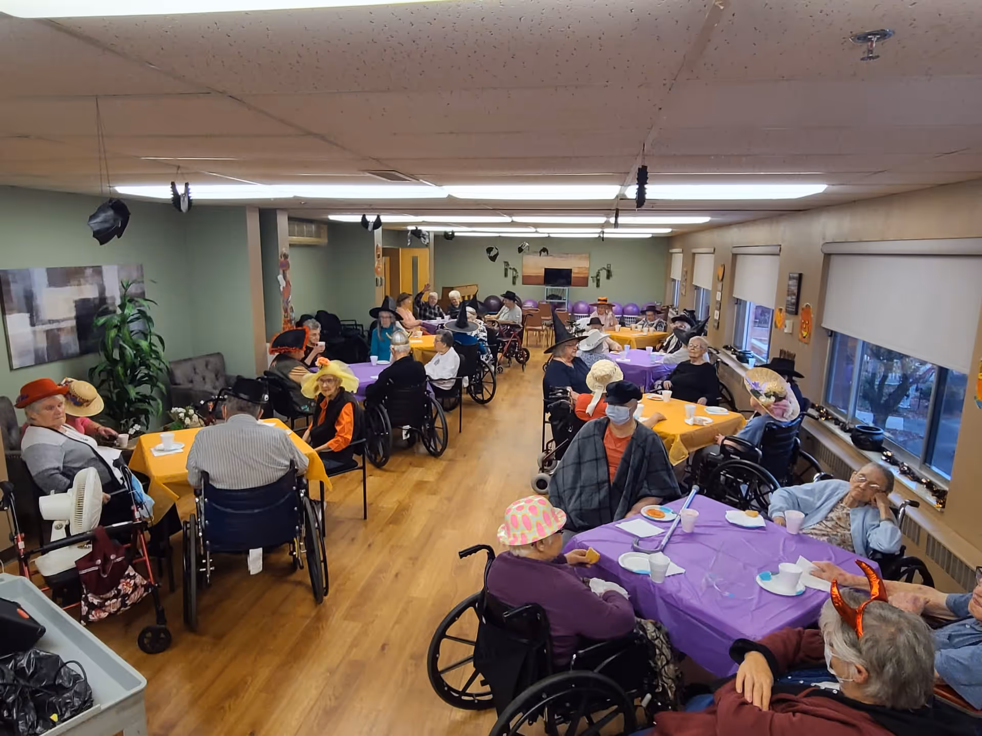 Seniors in wheelchairs wearing festive hats sit at decorated tables in a large communal dining/activity room.