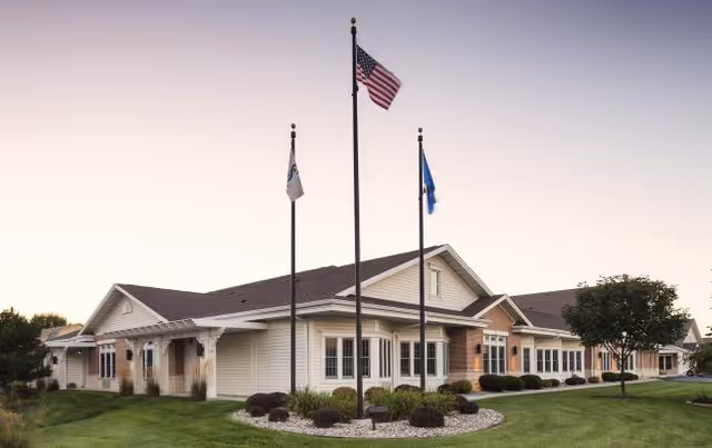 Single-story senior living facility building with three flagpoles and a manicured lawn at the entrance.