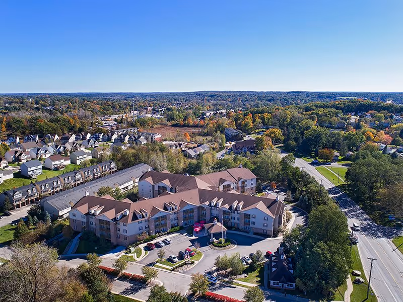 Aerial view of a large senior living facility with multiple connected buildings surrounded by trees and residential houses under a clear blue sky.