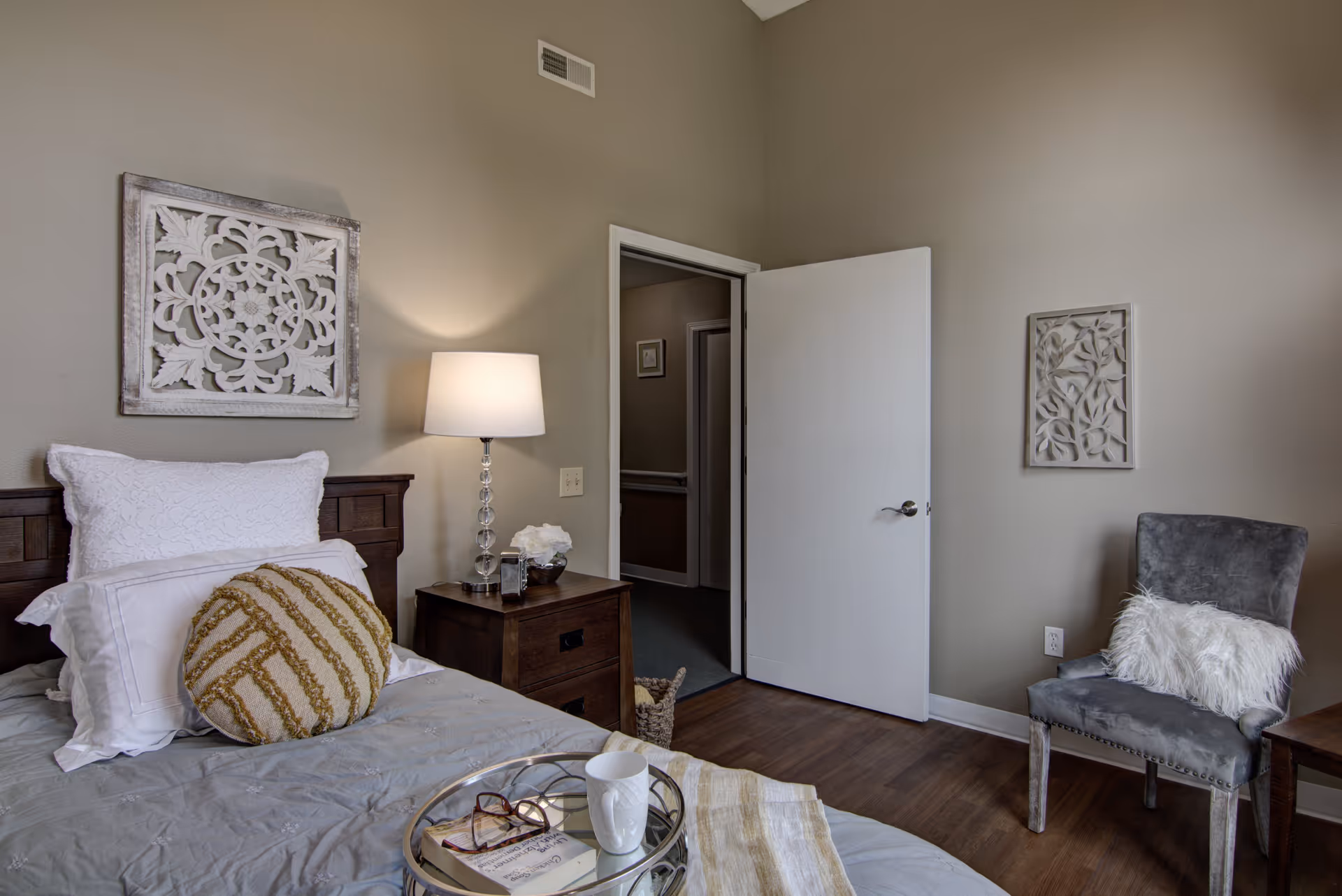 A cozy bedroom with a neatly made bed featuring white and beige pillows, a wooden nightstand with a lamp and decorative items, a gray upholstered chair with a white fluffy pillow, and two decorative wall hangings. The door to the room is open, showing a hallway outside.