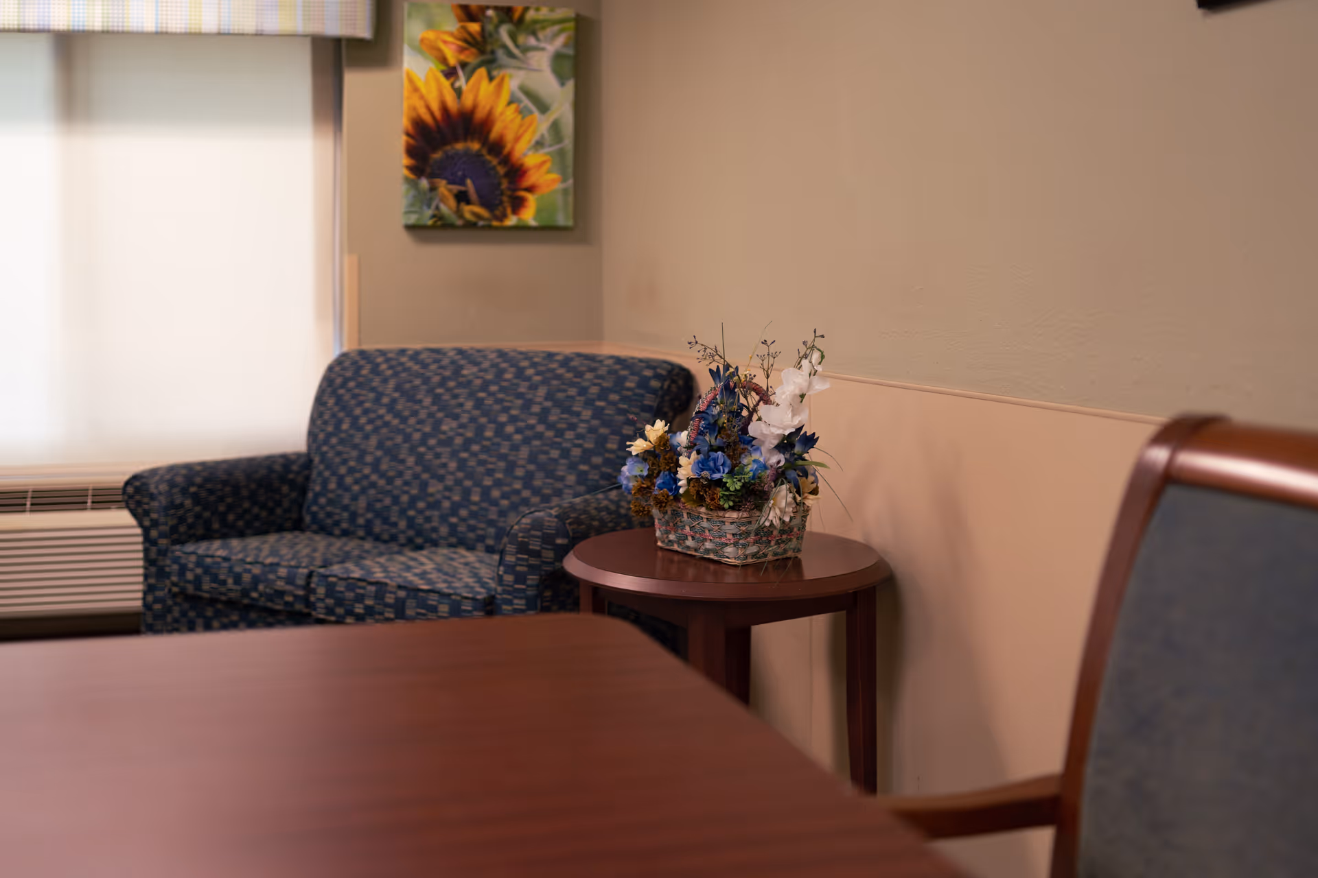 A cozy corner of a room featuring a blue patterned loveseat, a wooden side table with a floral arrangement in a basket, a wooden chair with blue upholstery, a window with a light-colored blind, and a sunflower painting on the wall.