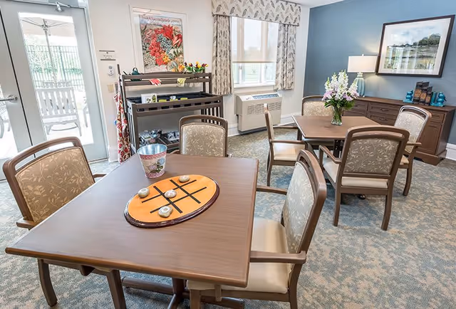 A bright and cozy common area in a senior living facility with two wooden tables, each surrounded by four cushioned chairs. One table has a tic-tac-toe game set on it. The room features a large window with patterned curtains, a sideboard with a lamp and decorative items, a floral painting on the wall, and a door leading to an outdoor patio with rocking chairs.