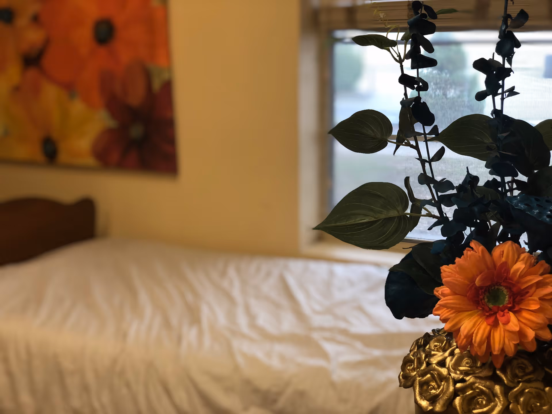 A close-up view of a decorative flower arrangement with orange flowers and green leaves in a golden vase, placed near a window. In the background, there is a bed with white bedding and a wall with a colorful floral painting.