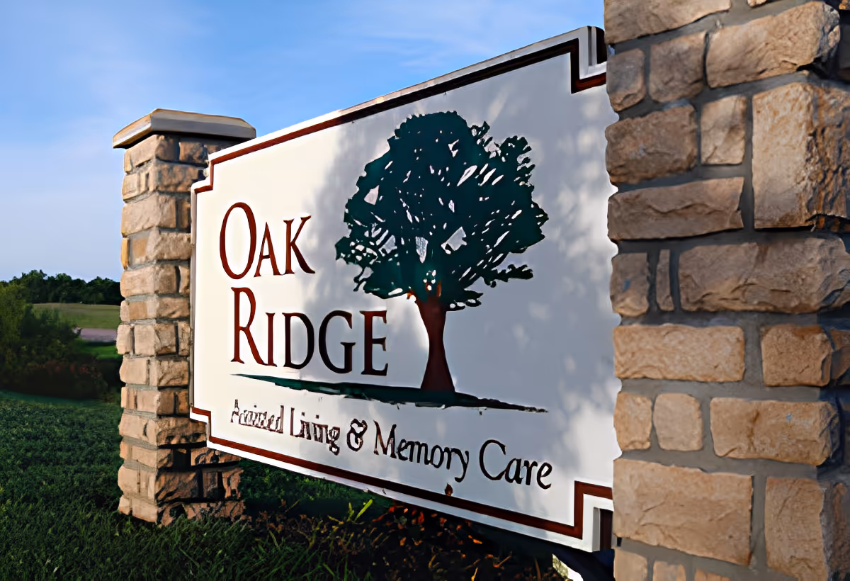 A large outdoor sign for Oak Ridge Assisted Living & Memory Care, mounted between two stone pillars with a green tree graphic and clear blue sky in the background.
