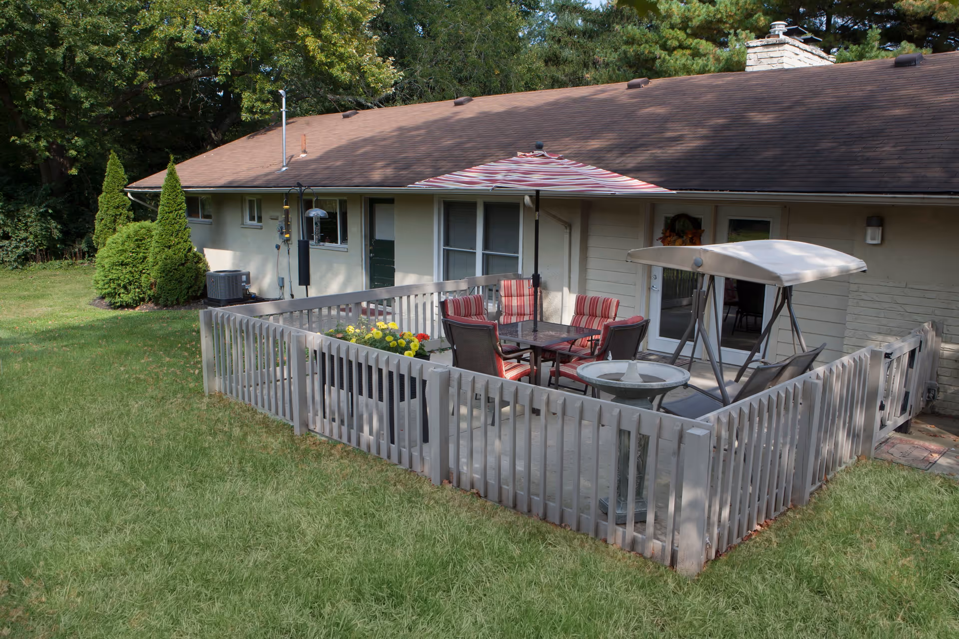 Outdoor fenced patio area attached to a single-story building with a brown roof. The patio contains a table with red and white striped chairs, a large umbrella, a swing with a canopy, and a planter with yellow flowers. The surrounding area has green grass and trees in the background.