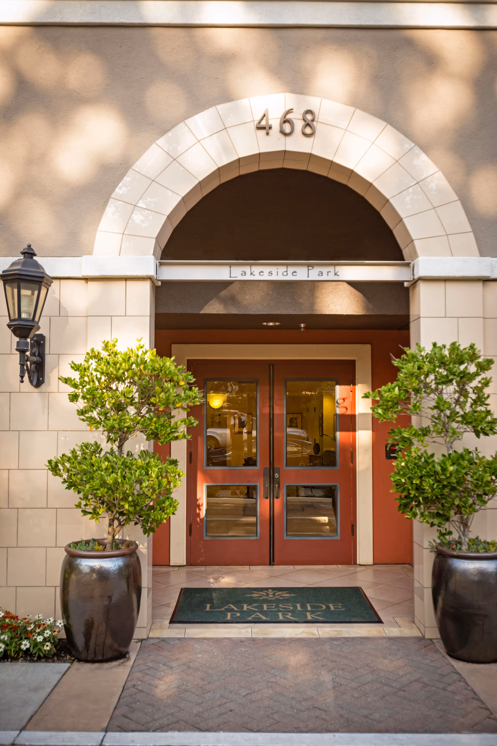 Entrance to a building with a beige tiled archway above double red doors. The number 468 is displayed above the arch, and a sign reading 'Lakeside Park' is above the doors. Two large potted plants flank the entrance, and a green doormat with the text 'Lakeside Park' is placed in front of the doors.
