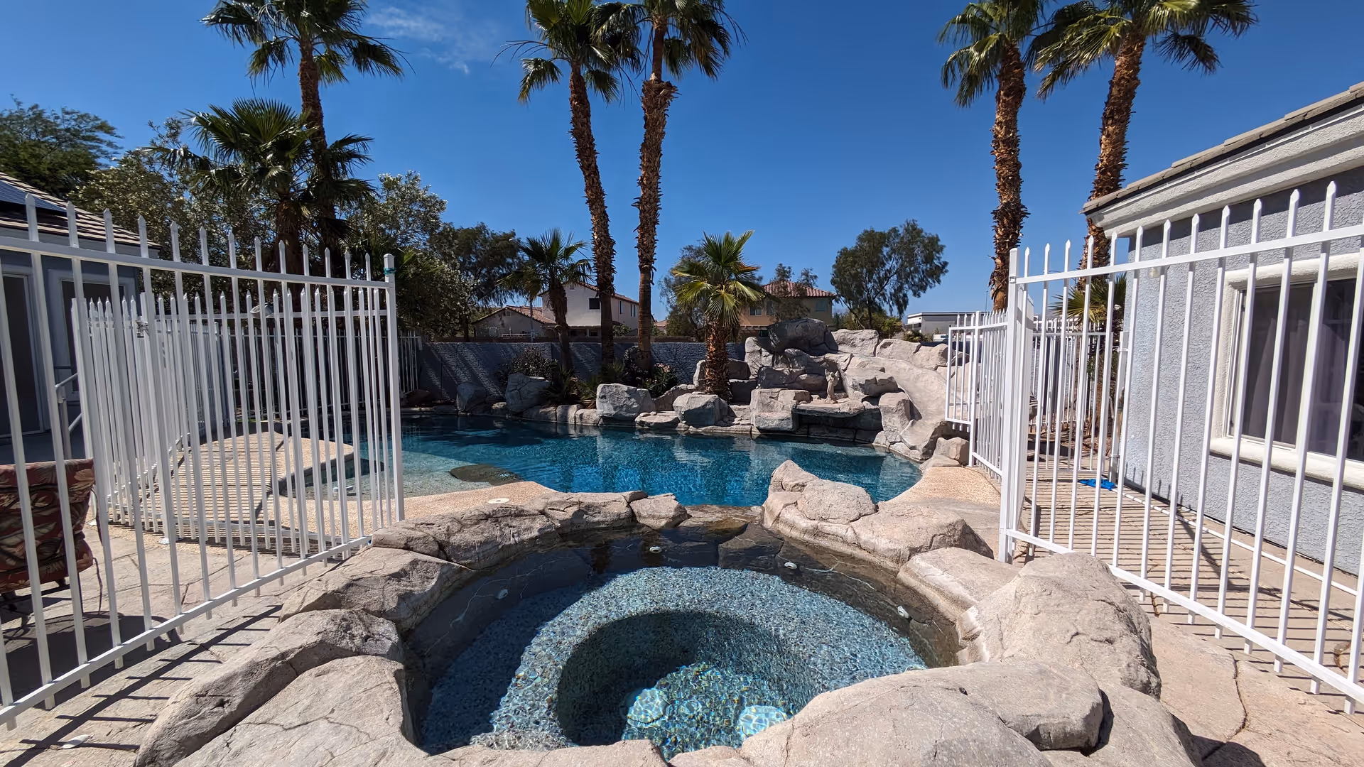 Outdoor pool area with a hot tub in the foreground surrounded by rock formations and a white metal fence. Palm trees and other greenery are visible around the pool under a clear blue sky.