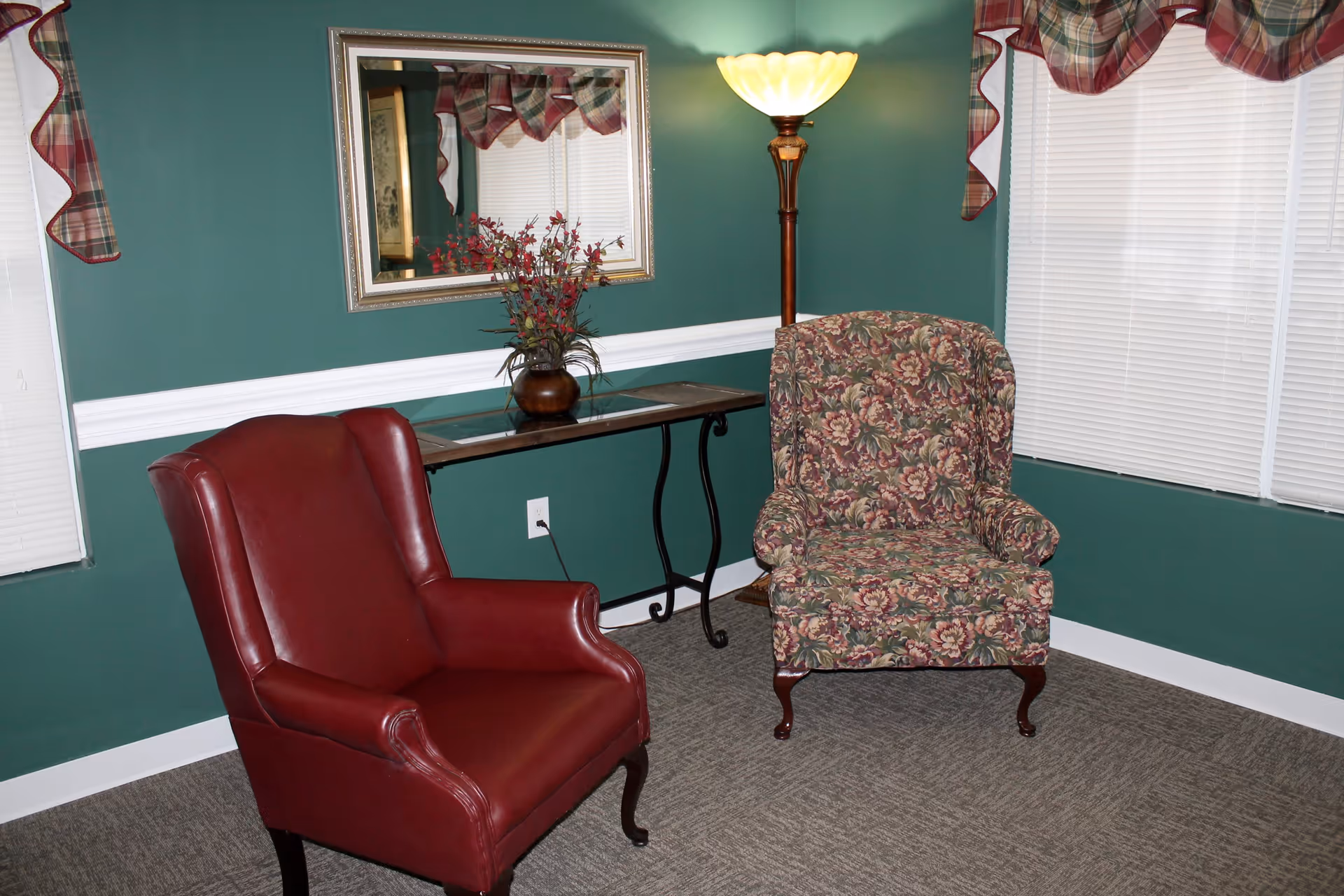 A cozy sitting area with two armchairs, one upholstered in red leather and the other in floral fabric, placed on a carpeted floor. Behind the chairs is a narrow table with a vase of flowers and a large mirror hanging on a green wall. A tall floor lamp with a yellowish shade is lit in the corner, and windows with white blinds and plaid valances are visible.