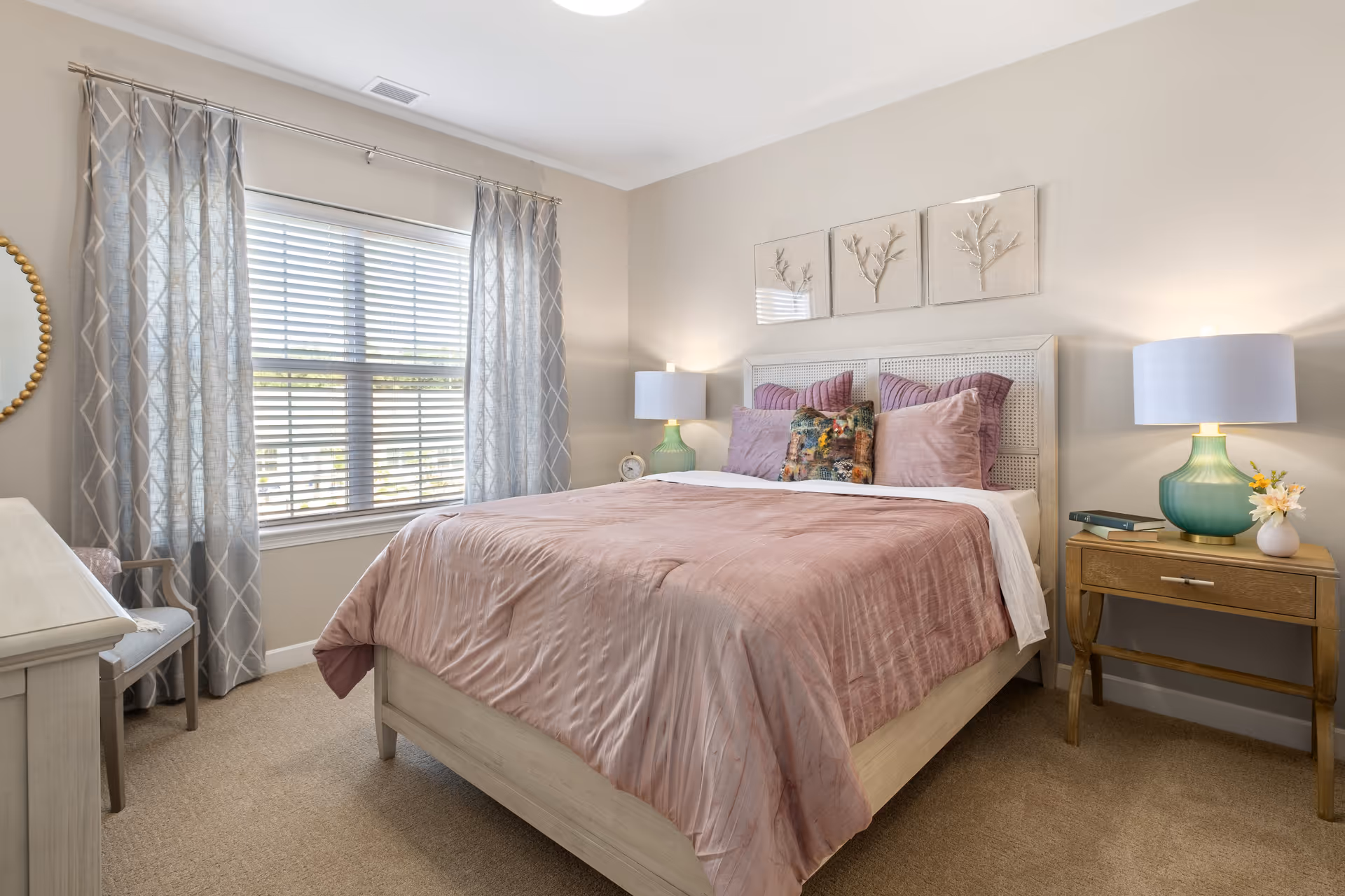 A cozy bedroom with a large bed covered in a soft pink comforter and multiple pillows. The bed has a light-colored wooden frame and a headboard with a woven design. On either side of the bed are wooden nightstands, each with a lamp featuring a green glass base and white shade. Above the bed are three decorative wall art pieces with a branch motif. A window with white blinds and gray patterned curtains lets in natural light. A chair and a dresser are partially visible in the room.