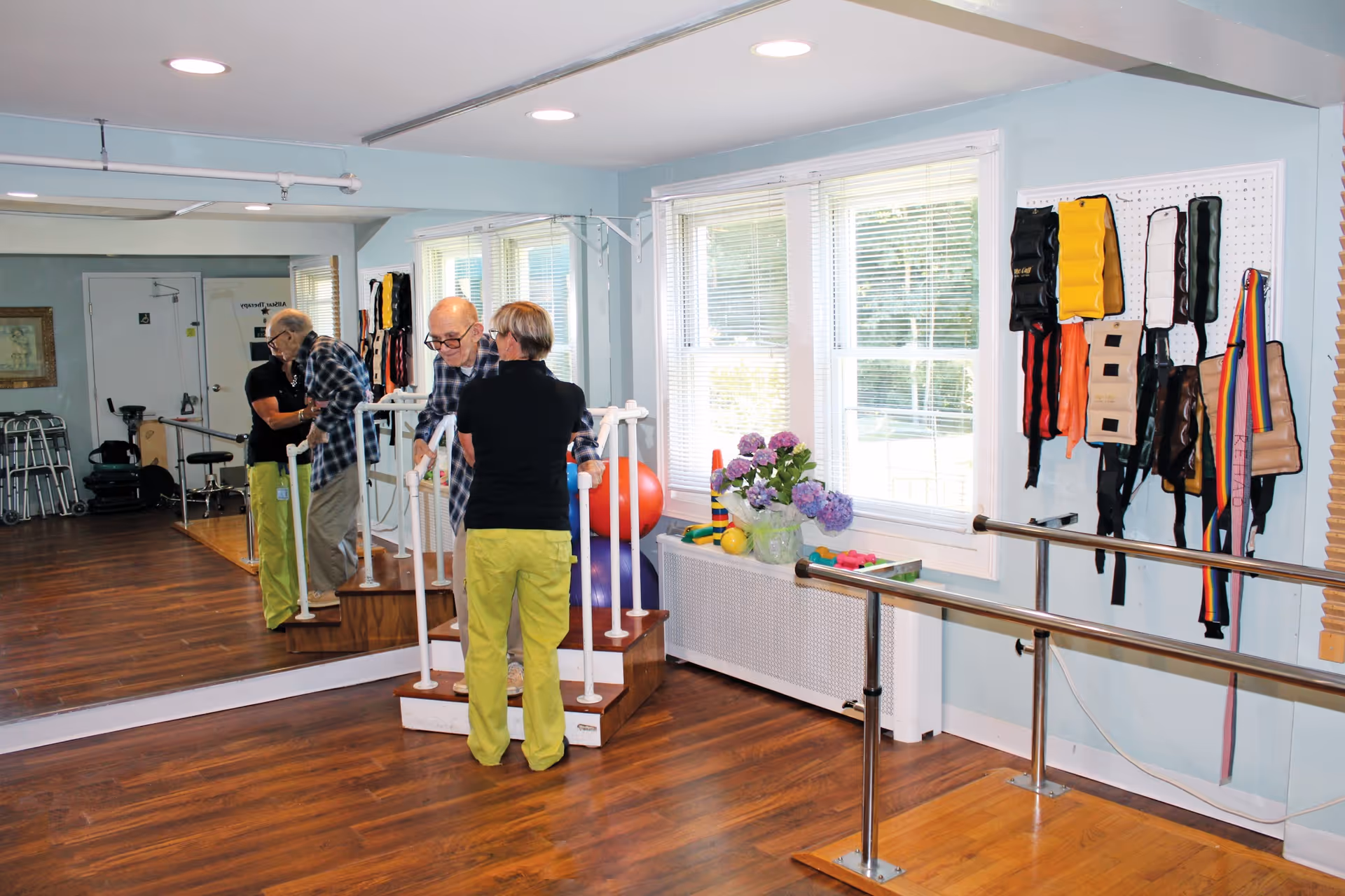 An elderly man is assisted by a caregiver as he practices walking up and down a small set of stairs with handrails in a rehabilitation or physical therapy room. The room has wooden floors, large windows with blinds, exercise equipment including therapy balls, and various weighted belts hanging on the wall. A large mirror covers one wall, reflecting the scene.