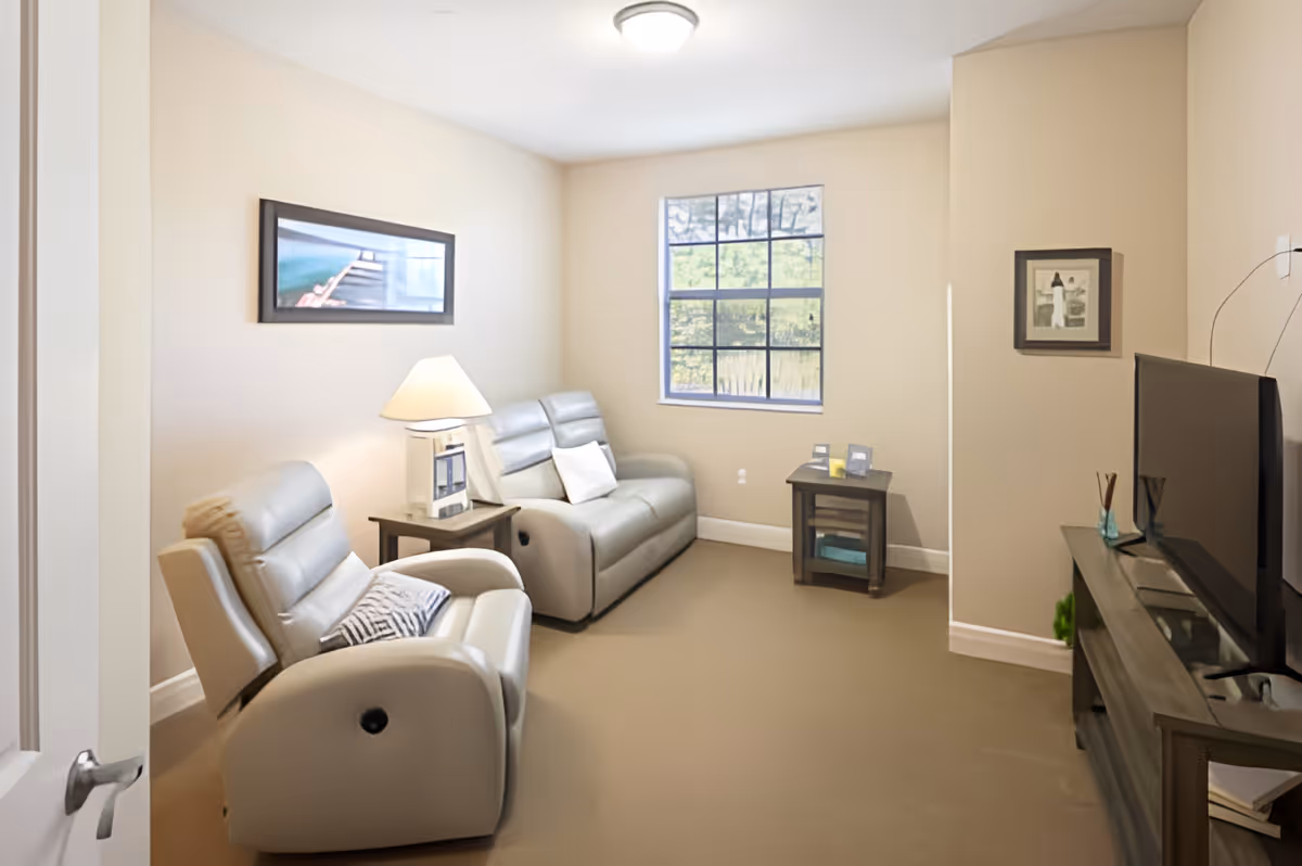A small living room with beige walls and carpet, featuring two light gray recliner chairs, a side table with a lamp, a window with a view of greenery outside, a framed picture on the wall, and a flat-screen TV on a wooden TV stand.