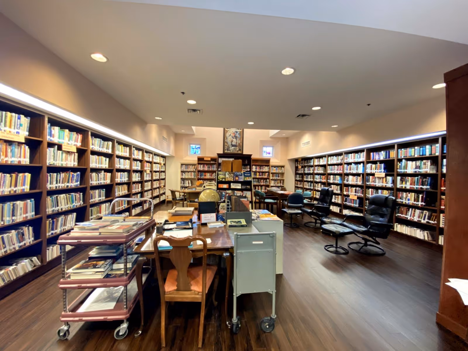Interior view of a library room with wooden bookshelves filled with books lining the walls. There are tables and chairs in the center, a book cart with books on the left, and several black leather chairs with footrests on the right. The room has recessed ceiling lights and small windows near the ceiling.