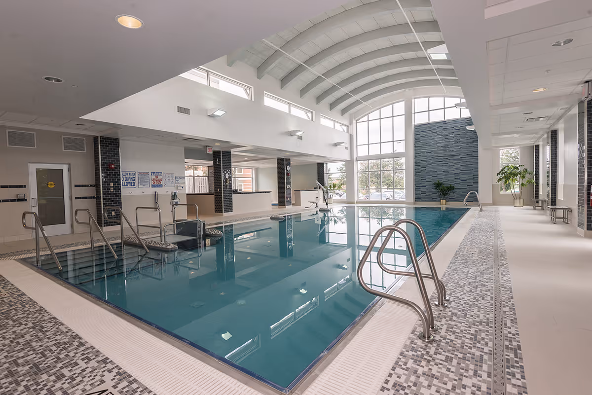 Indoor swimming pool area with clear blue water, metal handrails, tiled flooring, large windows letting in natural light, and potted plants along the side.