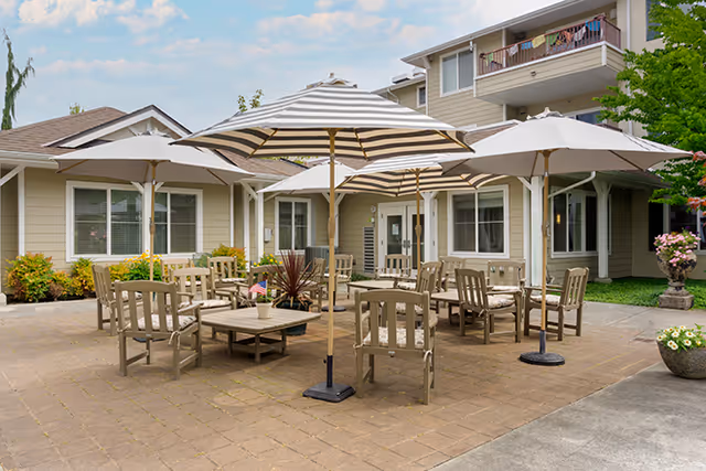 Outdoor patio area at Brookdale Stanwood with multiple wooden chairs and tables under large striped umbrellas, surrounded by a beige building with windows and balconies, and some greenery and potted plants.
