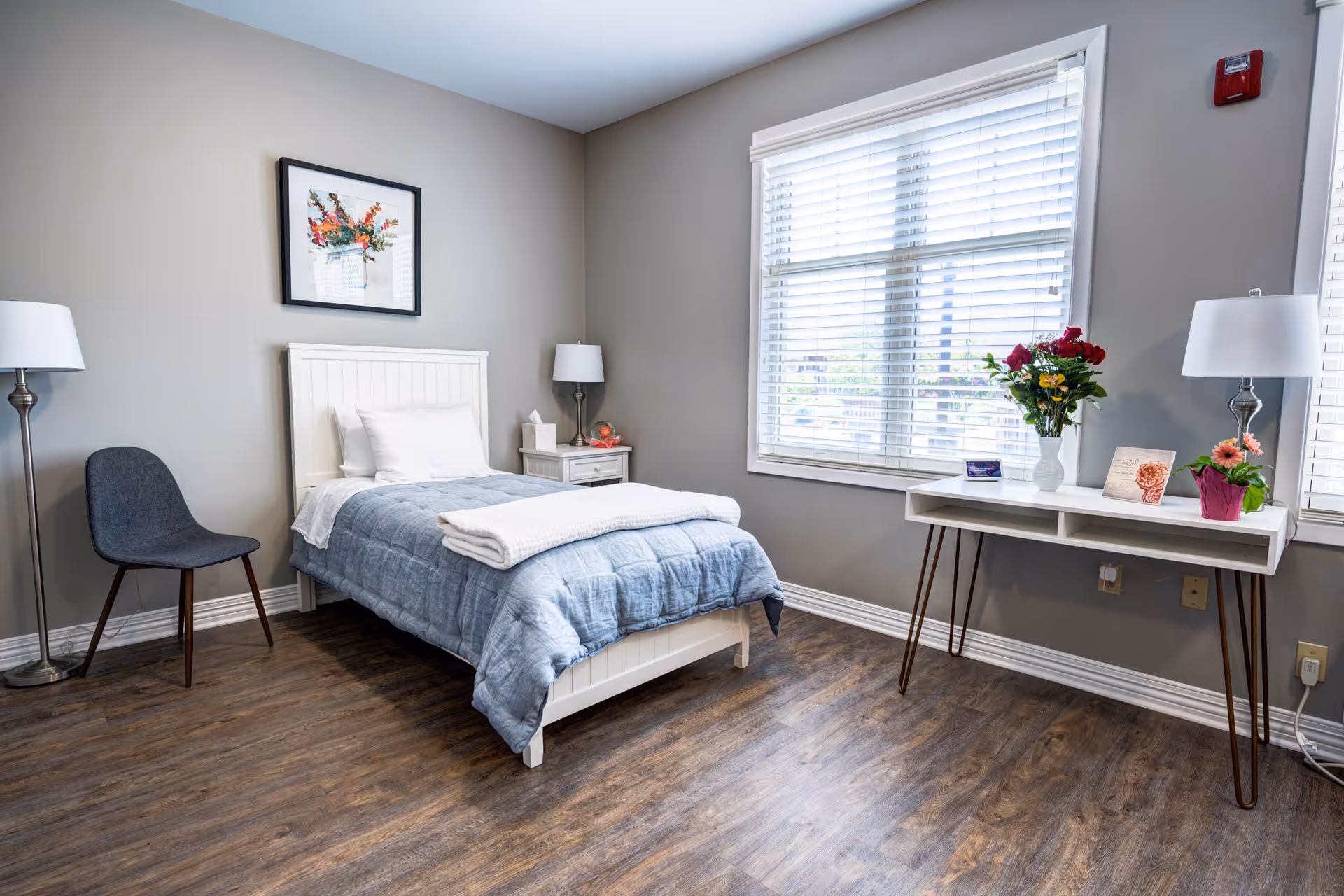 Bright, neatly furnished bedroom with a single bed, bedside table, desk with flowers, chair, and a window with blinds.