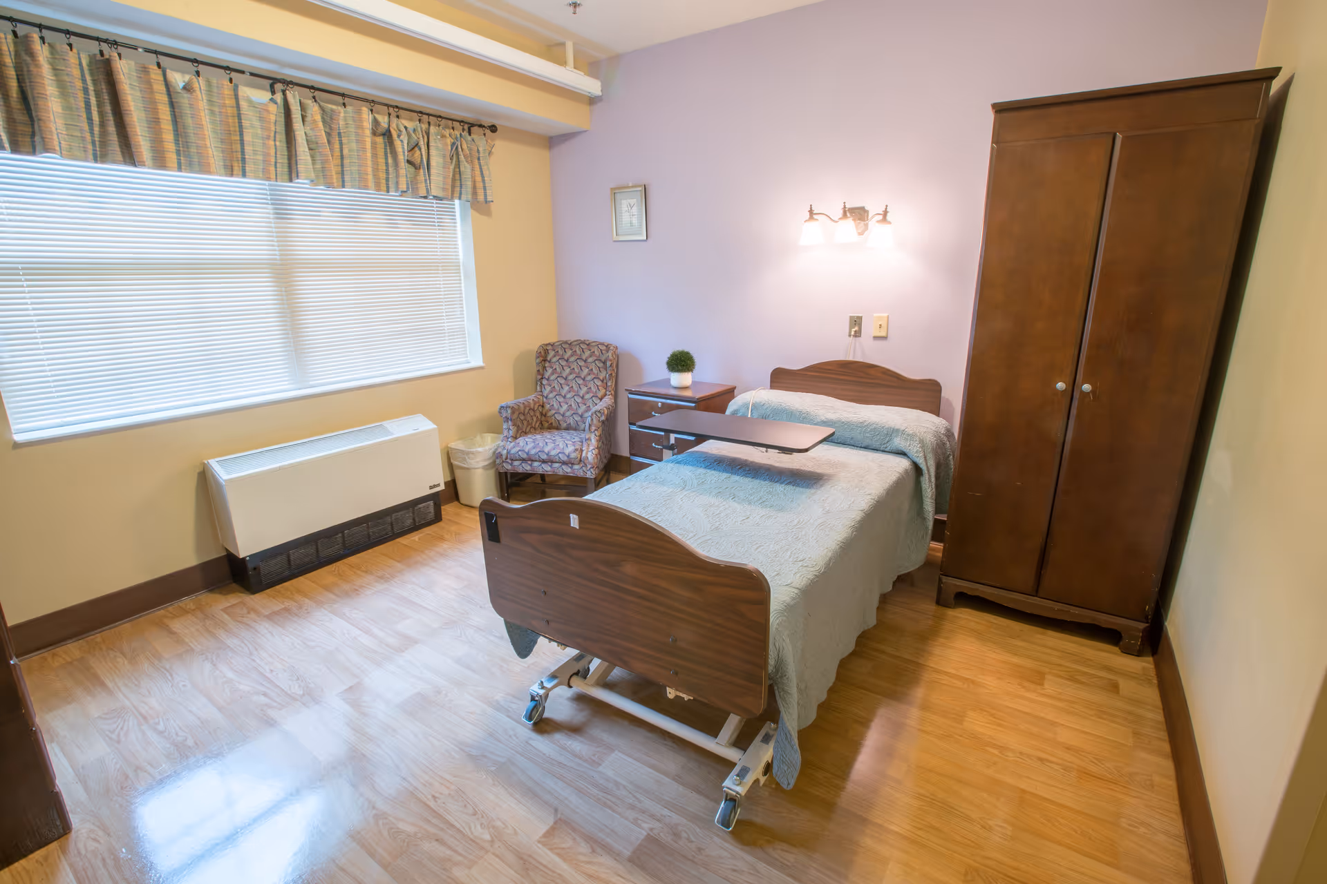A small bedroom in a senior living facility with a single hospital-style bed covered with a light blue quilt. Next to the bed is a wooden nightstand with a small potted plant and a patterned armchair. A wooden wardrobe stands against the wall. The room has light-colored walls, a large window with blinds and a valance, and wood flooring.