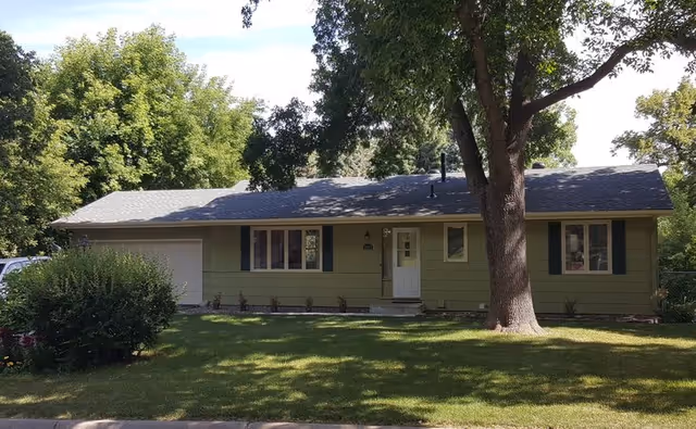 Single-story green house with a gray roof, white front door, and several windows, surrounded by green grass, trees, and bushes on a sunny day.