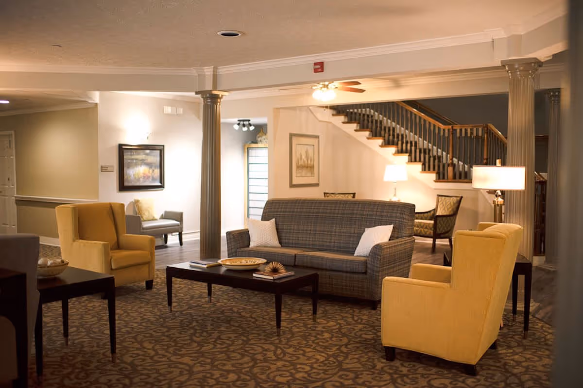 A cozy living room area in a senior living facility featuring a plaid sofa with two white pillows, two yellow armchairs, a dark wooden coffee table with decorative items, patterned carpet, and a staircase in the background. The room is softly lit with table lamps and ceiling lights, and there are framed pictures on the walls.