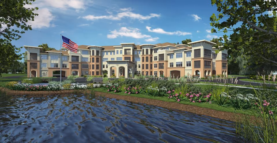 Exterior view of a large, multi-story senior living facility named Fleet Landing, with an American flag on a flagpole in front, surrounded by landscaped gardens and a pond in the foreground under a blue sky with scattered clouds.