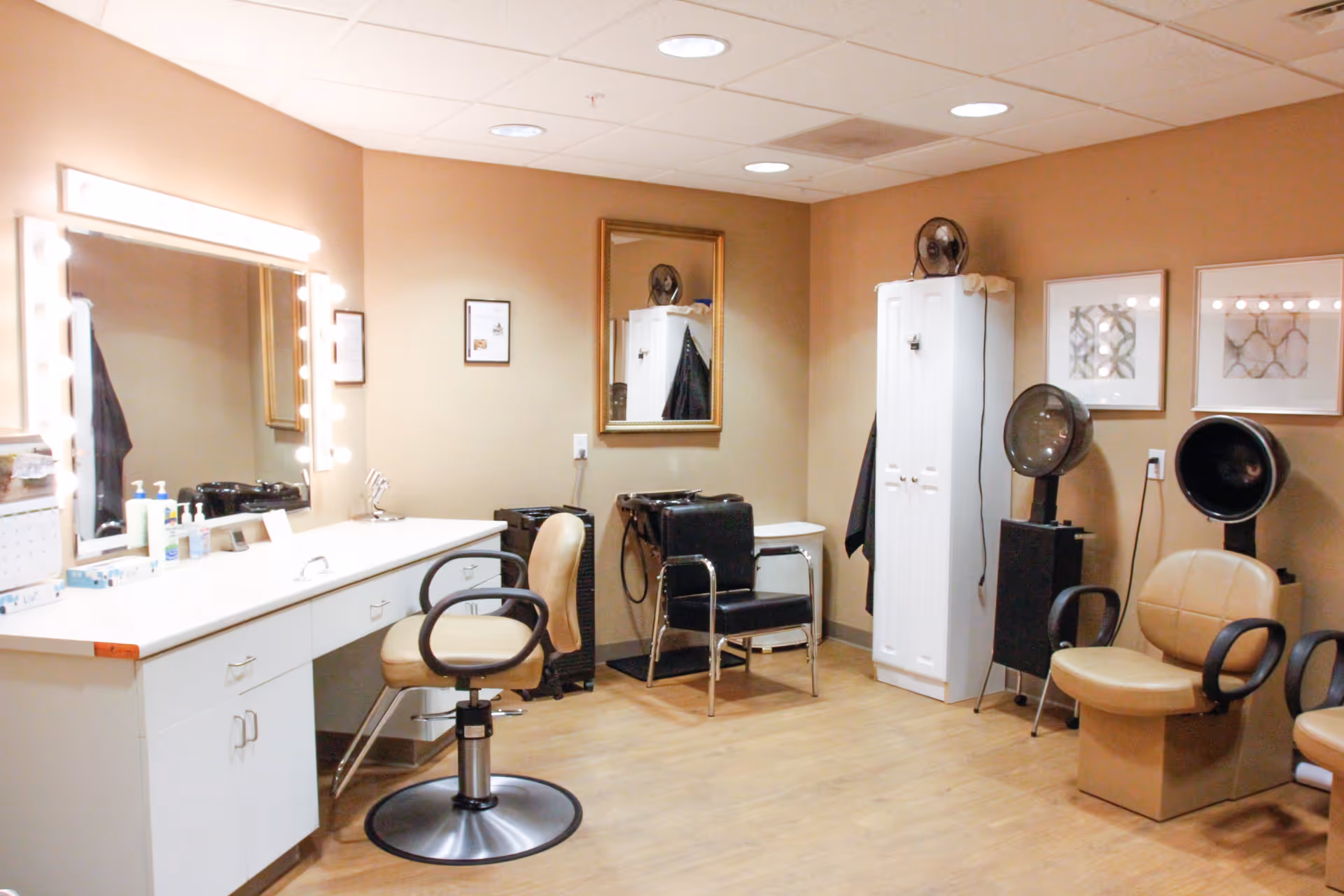 Interior view of a hair salon room with beige walls and wooden floor. The room features a large mirror with lights around it, a white counter with drawers, salon chairs, hair washing stations, and hair dryers. There are framed pictures on the wall and a white storage cabinet.