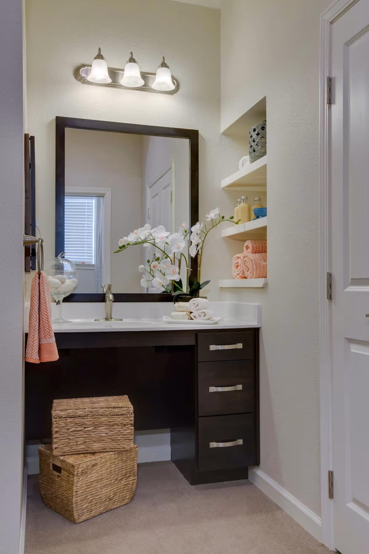 A bathroom vanity area with a large rectangular mirror framed in dark wood, a countertop with a sink and faucet, and decorative items including a white orchid plant, rolled towels, and a glass container with cotton balls. There are three light fixtures above the mirror, open shelves on the right holding folded towels and bottles, and two woven storage baskets on the floor beneath the vanity.