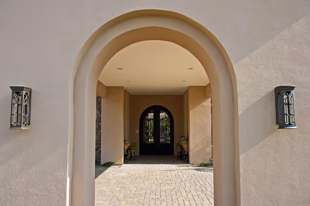 View through a beige stucco archway leading to a set of double black wrought iron doors with glass panels. The ground is paved with light-colored bricks, and there are two decorative wall lanterns mounted on either side of the archway.