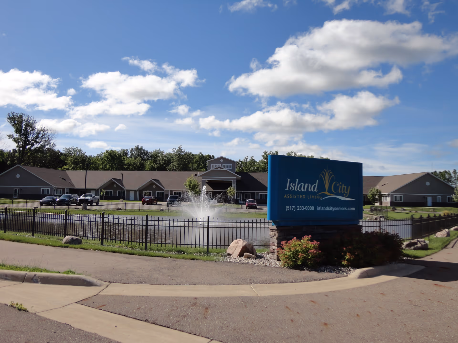 Exterior view of Island City Assisted Living facility with a large blue sign in the foreground displaying the facility name, phone number, and website. Behind the sign is a fenced pond with a water fountain, and a single-story building with multiple windows and a central entrance. The sky is blue with scattered clouds.