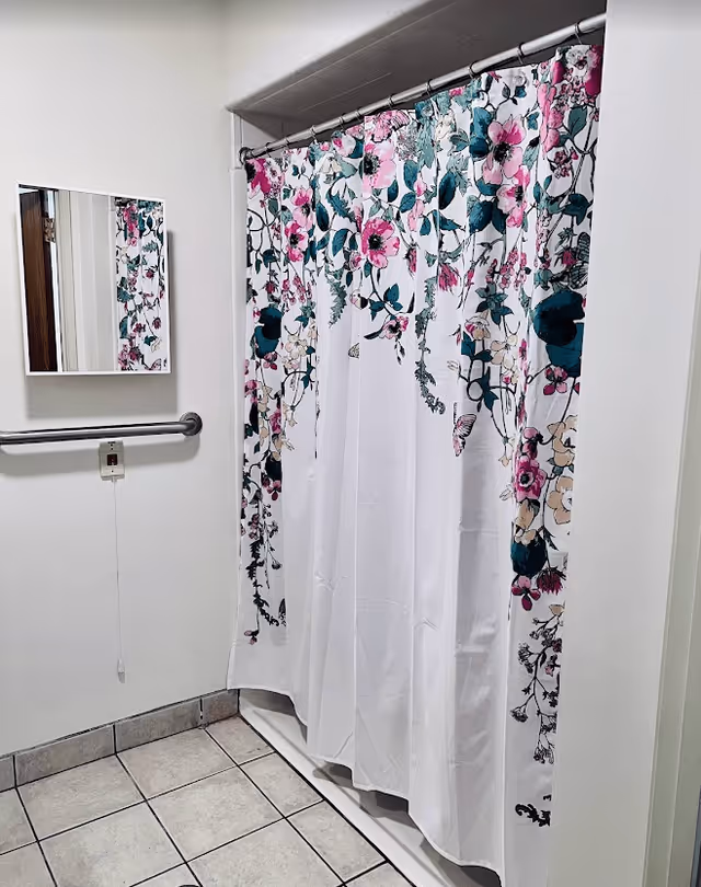 A bathroom with a floral shower curtain featuring pink, green, and beige flowers. There is a small rectangular mirror on the wall above a horizontal grab bar. The floor is tiled with light-colored tiles.