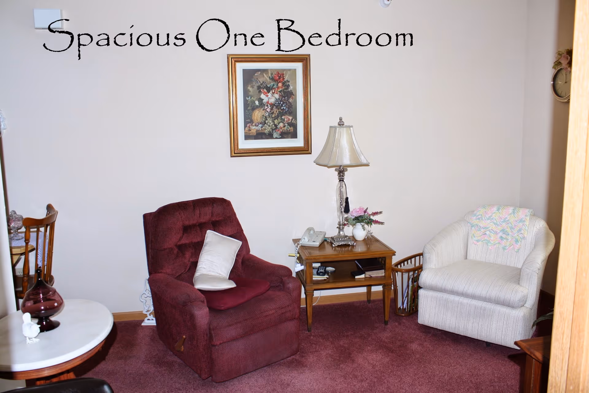 Cozy sitting area with a burgundy recliner, light armchair, side table with lamp and wall art in a carpeted room.