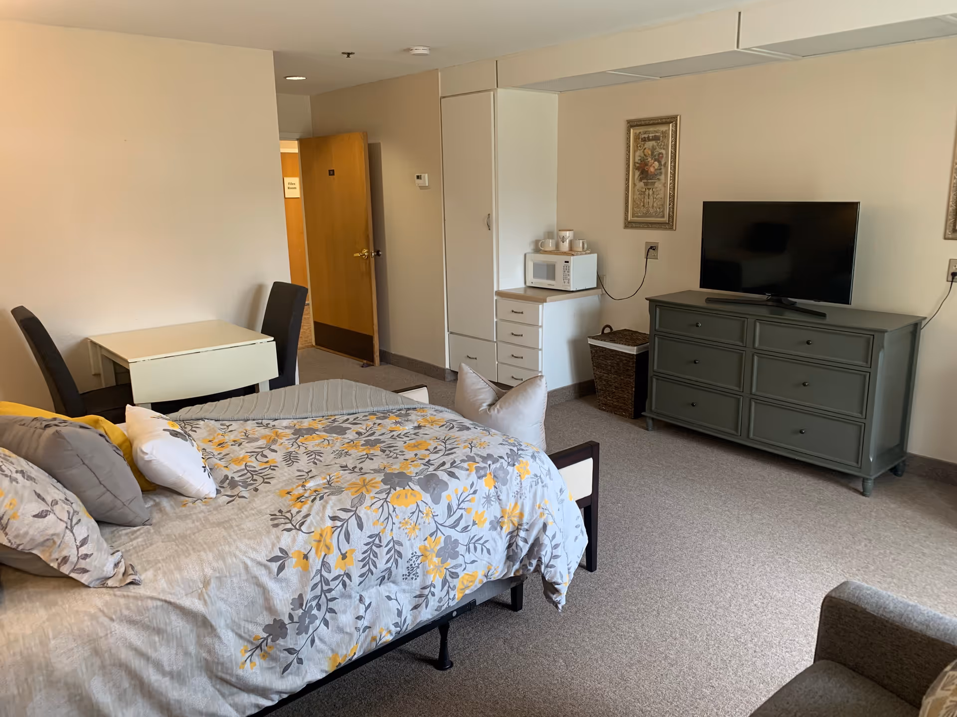 A cozy assisted living room with a bed covered in a floral patterned comforter in shades of gray and yellow. There is a small dining table with two chairs near the door, a green dresser with a flat-screen TV on top, and a white cabinet with a microwave and cups. The room has beige walls and carpeted floor.