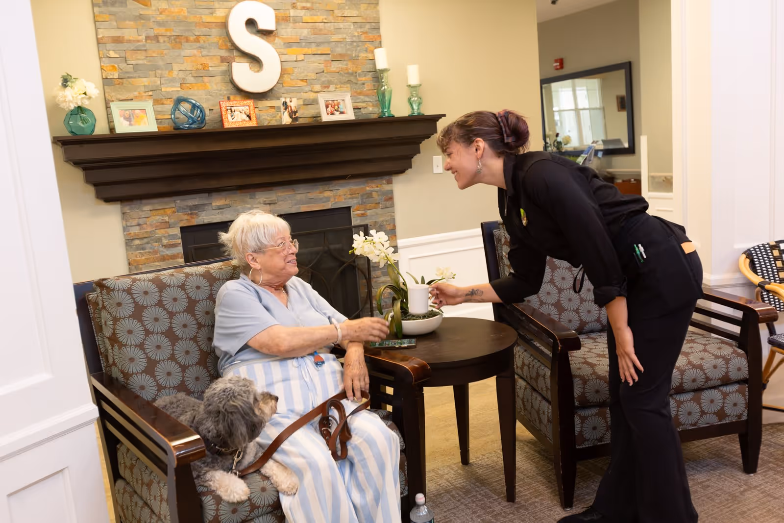 An elderly woman sitting in a patterned armchair with a small dog on her lap, smiling and reaching out to receive a cup from a caregiver who is leaning forward and smiling. They are in a cozy room with a stone fireplace, decorated mantel with photos and candles, and a large letter 'S' on the wall above the mantel.